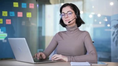 İş yerinde oturan gözlüklü ve kulaklıklı genç bayan ofis çalışanı portresi. Gülümseyen kadın müşteri hizmetleri temsilcisi modern ofis ortamında bilgisayarını kullanarak kameraya bakıyor.