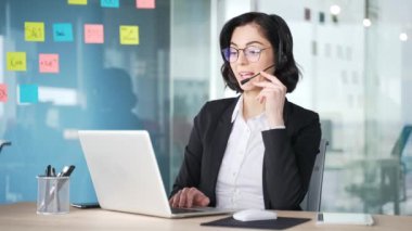 Müşteri hizmetleri temsilcisi ofis masasında dizüstü bilgisayarla çalışan kulaklık takıyor. Profesyonel kadın sohbete daldı, görevleri etkili bir şekilde yönetiyor. Kadın, destek veren müşteriler sunuyor