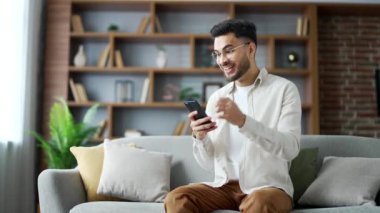 Mutlu yakışıklı adam evdeki oturma odasındaki koltukta otururken cep telefonundan harika haberler aldı. Gülümseyen sakallı erkek akıllı telefondan hoş bir mesaj okuyor ve kameraya bakarak başarısını kutluyor.