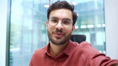 Gözlüklü, gülümseyen sakallı bir iş adamı, modern bir ofisteki bir telefonla video görüşmesinde konuşuyor. Yakışıklı yönetici kameraya el sallıyor. Bir müşteri veya iş ortağıyla çevrimiçi sohbet