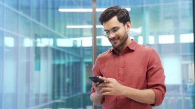 Gülümseyen işadamı iş yerinde dururken akıllı telefonu kullanıyor, tarıyor. Gözlüklü girişimci çevrimiçi sohbet ediyor, e- posta ya da yazıyı kontrol ediyor, istemciye kısa mesaj okuyor