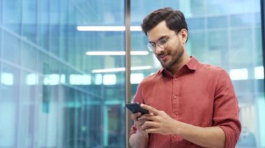 Mutlu, heyecanlı, yakışıklı iş adamı, modern bir iş ofisinde dururken telefonda harika haberler aldı. Gülümseyen girişimci akıllı telefondan olumlu bir mesaj okuyarak başarıyı kutluyor.