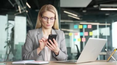 Ceketli düşünceli bir iş kadını iş yerindeki iş yerinde cep telefonu kullanıyor. Gözlüklü kadın yönetici uygulamada çalışır, internette sohbet eder ya da projeyi düşünür.