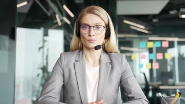 Webcam görüntüsü. Kablosuz kulaklıklı, kendine güvenen bir kadın müşteri temsilcisi iş yerinde kameraya bakarak görüntülü konuşma yapıyor. Çağrı merkezinin temsilcisi istemciye tavsiyede bulunur
