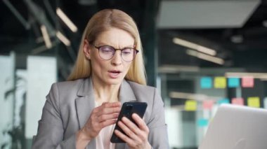 Şok olmuş ceketli iş kadını, iş yerinde masa başında otururken cep telefonundan kötü haberler okuyor. Endişeli kadın, akıllı telefondaki olumsuz mesaj yüzünden hayal kırıklığına uğradı. Kapat.
