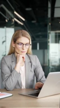 Takım elbiseli düşünceli bir iş kadını iş yerindeki laptopta çalışırken problem çözmeyi düşünüyor. Ciddi kadın çalışanlar bir projeyle meşgul. Dikey video
