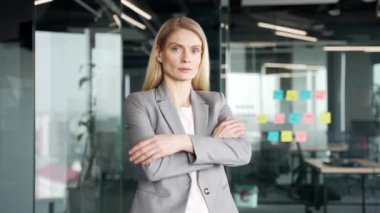 İş yerinde duran takım elbiseli bir iş kadınının portresi. Kollarını kavuşturup kameraya bakan ciddi sarışın kadın. Modern çalışma alanında poz veren başarılı kadın yöneticinin kafa fotoğrafı.