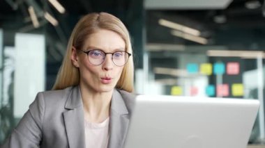 Ceketli mutlu iş kadını laptopta iyi haberler okuyor iş yerindeki masada oturuyor. Gülümseyen mutlu bir kadın, bilgisayardaki olumlu bir mesajla sevinir. Kapat.