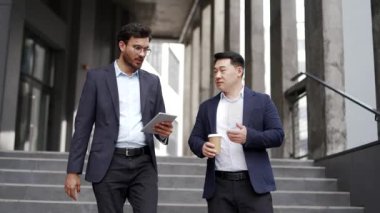 Modern ofis binasının önünde merdivenlerden inen iki işadamı iş fikirlerini tartışıyor. Resmi takım elbiseli meslektaşlar tablet, kahve, takım çalışması, işbirliği, profesyonel iletişim içerir