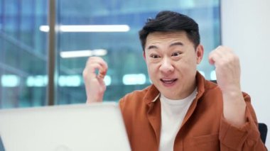 Mutlu Asyalı işadamı iş yerinde otururken dizüstü bilgisayarından harika haberler aldı. Gülümseyen şaşkın girişimci bilgisayardan olumlu bir mesaj okuyor. Kapat.