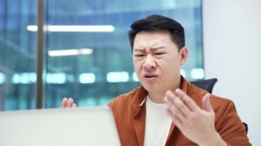 Endişeli Asyalı işadamı iş yerindeki masada otururken dizüstü bilgisayardan kötü haberler okuyor. Hüsrana uğramış işçiler bilgisayarda olumsuz bilgileri incelerken üzülüyorlar. Kapat.