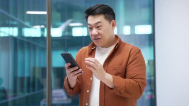 Mutlu Asyalı işadamı modern iş ofisinde dururken cep telefonundan harika haberler aldı. Gülümseyen şaşkın girişimci akıllı telefondan olumlu bir mesaj okuyor.