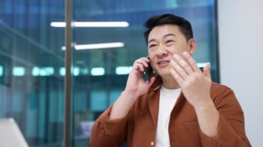 Modern ofisteki işyerinde otururken kendine güvenen Asyalı işadamı cep telefonuyla konuşuyor. Yönetici bir müşteriyle iletişim kurar ya da bir iş ortağıyla sohbet eder. Kapat.