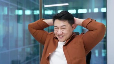 Gülümseyen Asyalı erkek profesyonel ofiste rahatlıyor, sandalyeye yaslanıyor. Moladan zevk alan iş adamı, modern çalışma alanından memnun ve kendinden emin. Erkek dinleniyor, ellerini başının arkasına koyuyor.