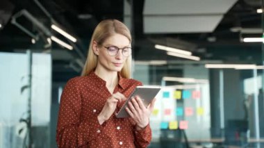 Ciddi düşünceli bir iş kadını iş yerinde dikilirken dijital tablet kullanıyor. Gözlüklü kadın yönetici uygulamada çalışır ya da proje hakkında düşünür, internette sohbet eder ya da internette gezinir