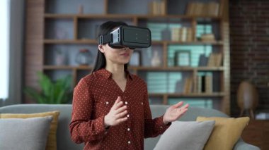 Video görüşmesi ve sanal gerçeklik simülatöründe sohbet için VR gözlüğü kullanan Asyalı genç bir kadın evdeki oturma odasında kanepede oturuyor. Kendinden emin bir kadın uzaktan iletişim kuruyor, online bir toplantısı var.