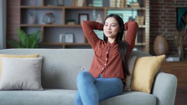 Genç gülümseyen Asyalı kadın, evinde oturma odasında kanepede otururken gözleri kapalı ve elleri başının arkasında dinleniyor. Sakin, mutlu, güzel bayan işten sonra dinleniyor, rahatlamış hissediyor.