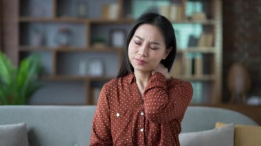 Evdeki oturma odasında kanepede otururken boynu ağrıyan genç ve yorgun bir Asyalı kadın. Gömlek giymiş üzgün bir kadın rahatsızlık hisseder, masaj yapar ve ağrıyan kaslarını ovuşturur, kendini gerer. Kapat.
