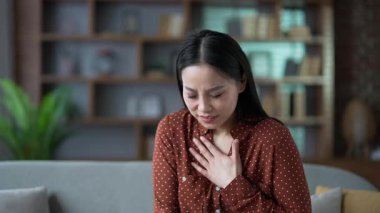 Genç Asyalı kadın evdeki oturma odasında kanepede otururken kalp krizi geçiriyor. Hasta kadın göğsündeki gerilimden muzdarip ve elleriyle hassas bir nokta tutuyor. Tıbbi yardıma ihtiyacı var. Kapat.