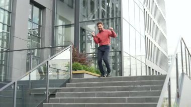 Kırmızı gömlekli iş adamı şehir merdivenlerinden iniyor güven ve enerji dolu bir şekilde dans ediyor. Heyecanlı erkek işçi zaferi kutluyor. Yakışıklı sakallı adam mutluluğu ifade ediyor. Başarı ve pozitif enerji.