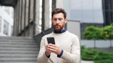Ciddi bir iş adamı bir ofis binasının yanında caddede dururken telefon kullanıyor. Yakışıklı sakallı erkek mesajlaşıyor, internette sohbet ediyor, internette geziniyor, akıllı telefondan mesajlar yazıyor.