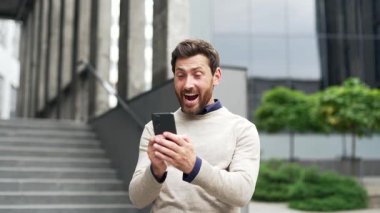 Dışarıda telefonları kontrol ederken başarıyı kutlayan mutlu sakallı erkek girişimci. Heyecanlı yakışıklı iş adamı harika haberler aldı. Ofis binasının yanında duruyor zafer ve memnuniyet sergiliyor