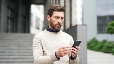 Hayal kırıklığına uğramış sakallı işadamı, bir iş yeri binasının yanında caddede dururken cep telefonundan kötü haberler okuyor. Üzgün adam akıllı telefonu kontrol ediyor, hayal kırıklığı ve endişe ifadesi gösteriyor.