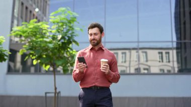 Kendine güveni olan kırmızı gömlekli bir girişimci modern şehir ofisinin dışında telefon kullanırken kahvenin tadını çıkarıyor. Sakallı profesyonel yürüyüş, akıllı telefon sohbetleri, internette gezinme, mesajlar yazıyor.