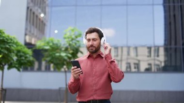 Şehirde kulaklık takmış ve telefon kullanan kendine güvenen bir iş adamı. Gülümserken müziği sever, modern yaşam tarzı dengesini somutlaştırır. Kırmızı gömlek, kentsel geçmiş, profesyonel canlılık.