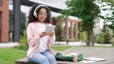 Öğrenmek için dijital tablet kullanan Afrikalı Amerikalı kız öğrenci üniversite binasının yanındaki bankta oturuyor. Kablosuz kulaklıklı genç siyahi kadın İnternet 'te ders veya internet sitesi izliyor