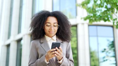 Ofis binasının dışında telefonla duran kendine güvenen bir iş kadını. Kadın profesyonel bağlantıda kalıyor ve akıllı telefon üzerinde çalışıyor. Modern iş ve teknolojinin temsili