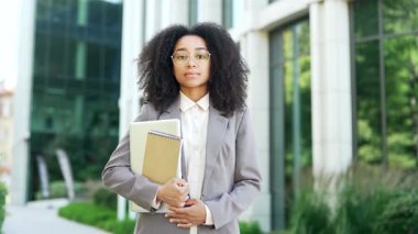 Açık havada elinde belgelerle duran, kendinden emin, ciddi bir Afrikalı Amerikalı iş kadınının portresi. Profesyonel ortam, arka planda modern ofis binası. Kariyerine odaklan. Öğretmenin vesikalığı.