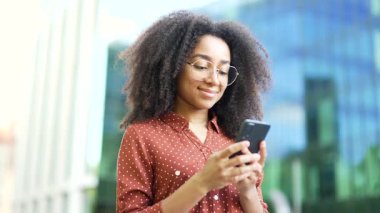 Afro-Amerikalı kadın açık havada akıllı telefon kullanıyor ve gülümsüyor. Gözlüklü ve günlük kıyafetlerle telefonu elinde tutan kişi. Mutlu zenci kadın profesyonel mesajları kontrol ediyor. Kapat.