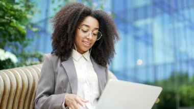 Gözlüklü, kendine güvenen siyahi kadın bilgisayarında uzaktan kumandalı video toplantısı yapıyor. Bilgisayarda çalışan Afrikalı Amerikalı iş kadını bir ofis binasının yanındaki bankta oturuyor. Kapat.