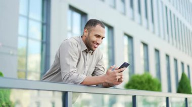 Yakışıklı adam internette sohbet ediyor. Sokaklara mesajlar yazıyor. Akıllı telefon kullanan bir erkek, sosyal medya ağlarına yazıyor ve mesajlar gönderiyor. Ofis binasının arka planında duruyor.