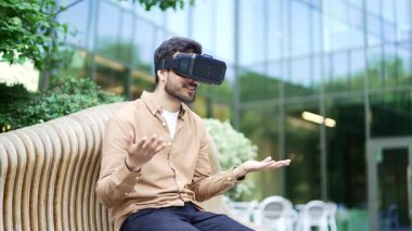 Başarılı bir iş adamı, sanal gerçeklik simülatöründe sanal gerçeklik simülatöründe video görüşmesi için VR gözlüklerini kullanıyor ve bir iş yeri binasının yakınındaki bir bankta otururken sohbet ediyor. Toplantı toplantısı uzaktan yapılıyor.
