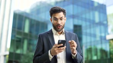 Takım elbiseli mutlu iş adamı, iş yerinin yakınındaki bir sokakta beklerken telefonda harika haberler okuyarak başarıyı kutluyor. Gülümseyen adam neşeli bir mesaj aldı, evet işareti yaptı.