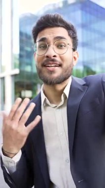POV dikey video. Takım elbiseli gülümseyen işadamı, bir iş yeri binasının yakınındaki bir sokakta cep telefonuyla konuşuyor. Çevrimiçi iletişim kuran mutlu işçi kameraya bakıyor.