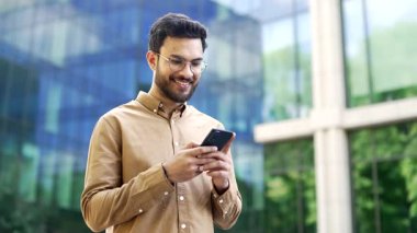 Modern cam ofis binasının yanında duran kendine güvenen işadamı akıllı telefonu elinde tutuyor. Gözlüklü adam, girişimci, işçi, sahibi veya yöneticisi dışarıda cep telefonu kullanıyor. Kapat.