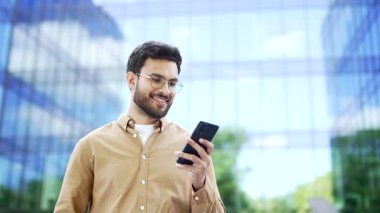 Modern cam ofis binasının yanında duran mutlu iş adamı akıllı telefonu elinde tutuyor. Gözlüklü sakallı adam, girişimci, işçi, sahibi ya da yöneticisi dışarıdaki cep telefonuna bakıyor.