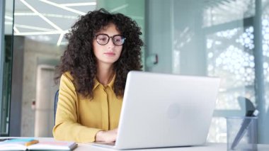 Modern iş ofisindeki iş yerinde dizüstü bilgisayarı başında otururken problem çözmeyi düşünen düşünceli genç iş kadını. Gözlüklü ciddi bir kadın bilgisayarda bir proje üzerinde çalışıyor.