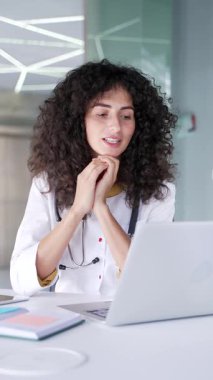 Modern hastane kliniğinde dizüstü bilgisayarla konuşan beyaz önlüklü genç bayan doktor. Tıp doktoru ofisteki bir iş yerinde masa başında oturan bir hastaya danışıyor. Dikey