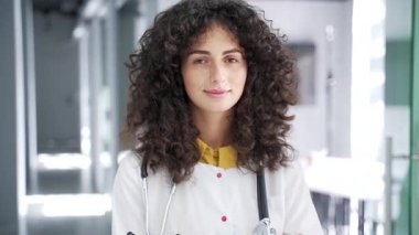 Modern hastane kliniğinde kameraya bakan beyaz önlüklü mutlu kadın doktor portresi. Yetişkin pozitif esmer kadın doktor ofiste poz veriyor. Kapat.