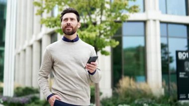 Modern ofis binasının dışında yürüyen gülümseyen işadamı iletişim için akıllı telefonu kullanıyor. Günlük giysiler içinde profesyonel bir erkek. Şehir hayatından zevk alıyor, mesajları kontrol ediyor ve bağlantıda kalıyor.