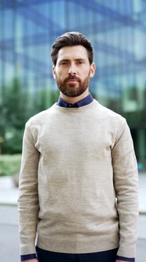 Dikey video. Şehrin caddesinde duran kendine güvenen güler yüzlü iş adamı. Gündelik kıyafetlerle şehir ortamında olgun bir erkek profesyonel. Modern ofis binasının yakınındaki açık hava yetişkin girişimcisi.