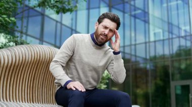 Başı ağrıyan bir iş adamı şehir ortamında bankta oturuyor, günlük kazak giyiyor, rahatsız edici bir yüz ifadesi sergiliyor. İşçi elleriyle şakaklarına masaj yapıyor. Çok yorgun.