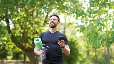 Sporcu, yoga minderi ve su şişesiyle parkta yürürken spor haberleri için telefona bakar. Gülümseyen sakallı sporcu antrenmandan dönüyor. Aktif yaşam tarzı, sağlıklı rutin