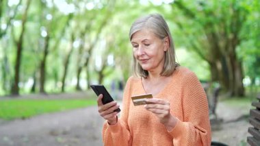 Açık hava bankında oturan olgun bir kadın akıllı telefon ve kredi kartı kullanıyor. Gülümsüyor ve sakin park ortamında dijital bankacılık veya çevrimiçi alışveriş yapıyor. Modern yaşam tarzı ve teknoloji