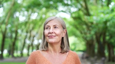 Şehir parkında kapalı gözlerle rahatlayan mutlu olgun bir kadın. Gri saçlı yaşlı kadın derin derin nefes alıyor ve doğada olmaktan hoşlanıyor. Dinginlik, rahatlama, açık havada farkındalık. Kapat.