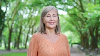 Parktaki parlak yeşil ağaçlarda duran mutlu yaşlı kadın portresi. Kadın kameraya sıcak bir şekilde bakıyor, pozitif yaşam tarzı ve doğadaki neşe. Aktif emeklilik ve açık hava eğlencesi. Kapat.